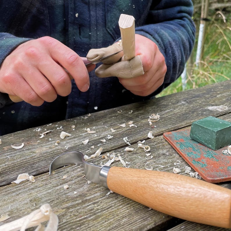 May include: A wood carving scene with a carving knife and a small wooden piece being worked on. The knife has a curved blade and a wooden handle. Wood shavings are scattered on the wooden table. A green block of wax and a small cutting board are also visible.