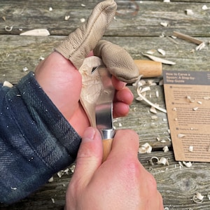 May include: A person carving a wooden spoon with a small knife. The spoon is partially carved and the person is wearing leather finger guards. The text "How to Carve a Spoon. A Step-by-Step Guide" is visible on a piece of paper in the background.