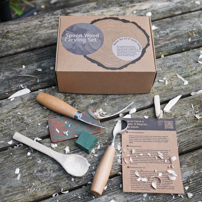 May include: A spoon wood carving set with a box, tools, and a step-by-step guide. The box is brown with the text "Spoon Wood Carving Set". The tools include a knife, a gouge, a spoon, and a block of wood. Wood shavings are scattered around the tools.