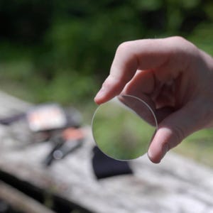 Op de afbeelding: Een heldere, cirkelvormige lens wordt tussen vingers gehouden tegen een vage groene achtergrond. De lens reflecteert de omgeving. De foto is buiten gemaakt, met natuurlijk licht.