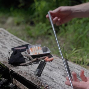 Op de afbeelding: Een zilverkleurig, opvouwbaar metalen rietje wordt vastgehouden voor een rustiek houten oppervlak. Andere kampeeruitrusting is zichtbaar, waaronder een kleine zwarte pouch, een metalen doos met lucifers en een klein mes. De achtergrond is wazig.