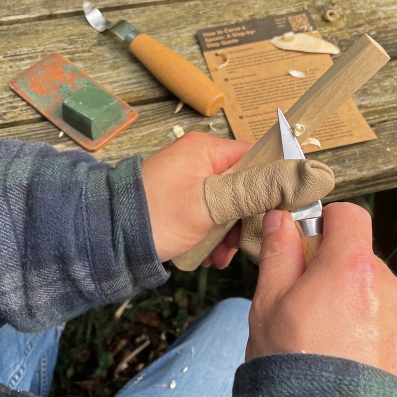 May include: A person is using a small knife to carve a piece of wood. They are wearing a leather finger guard to protect their thumb. The wood is light brown and the knife has a wooden handle. The text on the paper says "How to Carve a Spoon: A Step-by-Step Guide".