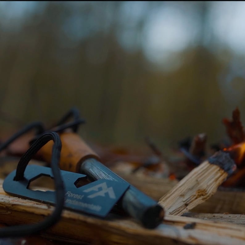 Puede incluir: Un iniciador de fuego negro con un mango de cuero marr&oacute;n y un percutor de metal. El iniciador de fuego est&aacute; sobre un trozo de madera con un peque&ntilde;o fuego ardiendo en el fondo. El iniciador de fuego tiene el texto "Forest Fundamentals" en &eacute;l.