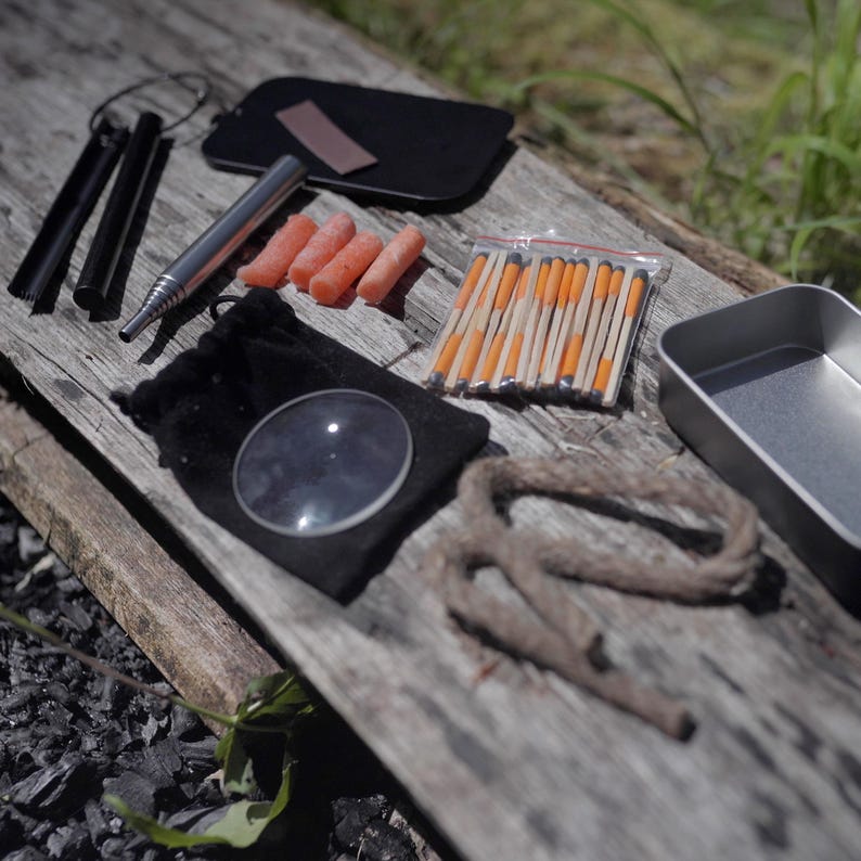 Op de afbeelding: Een survival kit met diverse gereedschappen voor gebruik buitenshuis. Inclusief een vergrootglas, lucifers, een metalen bakje, een vuurstarter, een klein zakje, een touw en een metalen buis. De items zijn gerangschikt op een verweerd houten oppervlak.