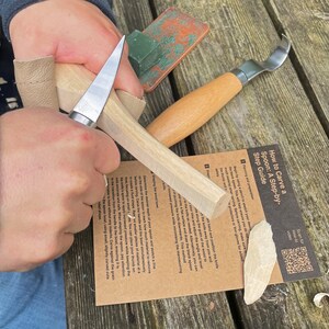 May include: A person's hands are carving a wooden spoon with a knife. The spoon is being carved from a block of wood. The image includes instructions on how to carve a spoon, with a QR code for a how-to video.