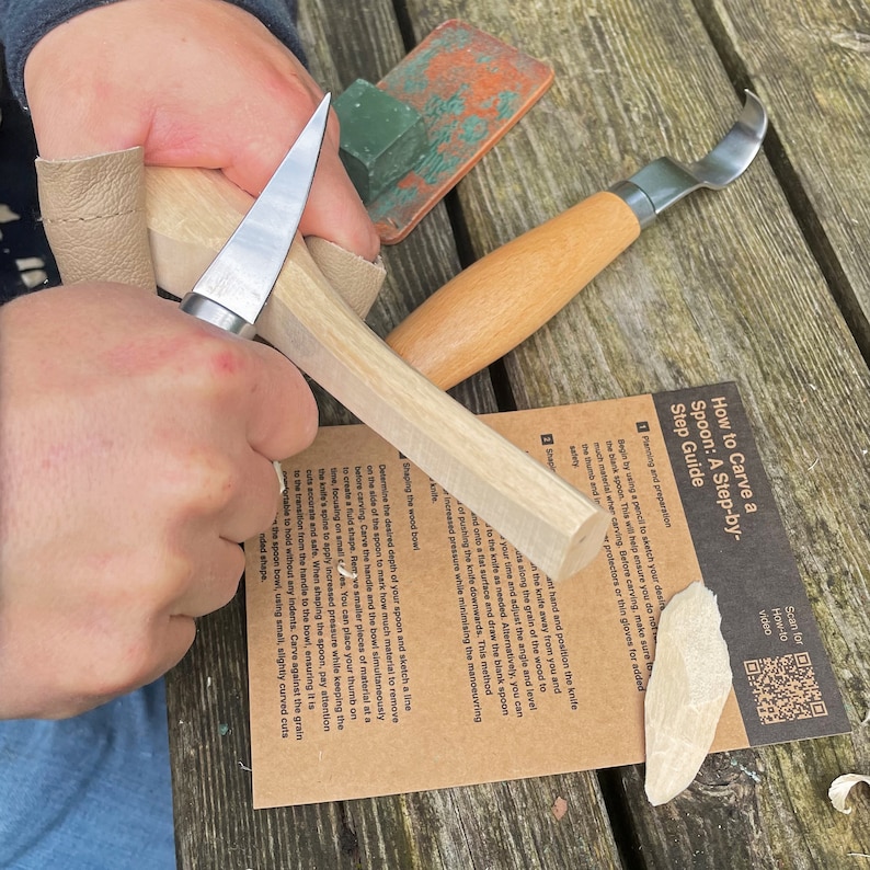 May include: A person's hands are carving a wooden spoon with a knife. The spoon is being carved from a block of wood. The image includes instructions on how to carve a spoon, with a QR code for a how-to video.