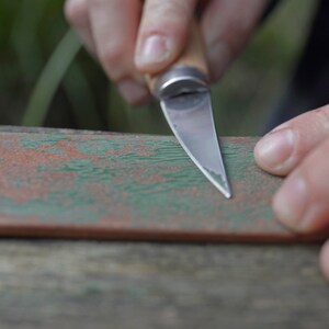 May include: A person sharpening a small knife on a piece of wood with green and brown paint. The knife has a wooden handle and a silver blade.