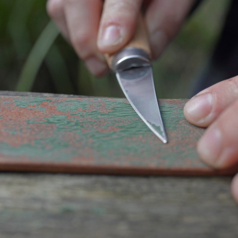 May include: A person sharpening a small knife on a piece of wood with green and brown paint. The knife has a wooden handle and a silver blade.
