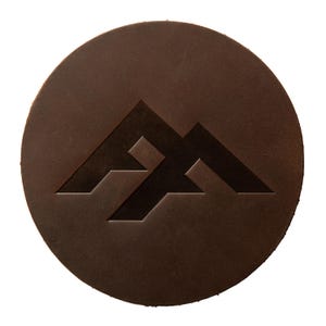 Könnte beinhalten: Ein runder Untersetzer aus dunkelbraunem Leder mit einem eingeprägten geometrischen Logo in der Mitte. Das Logo besteht aus zwei sich überschneidenden, kantigen Formen, die eine bergähnliche Silhouette bilden. Das Leder hat eine glatte, matte Oberfläche mit sichtbaren natürlichen Maserungen.