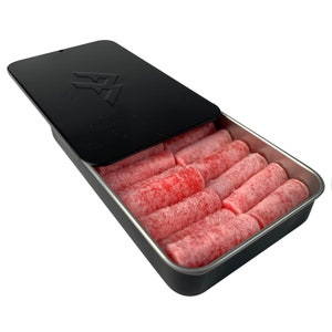 Puede incluir: Una caja metálica negra con tapa parcialmente abierta, que revela filas de caramelos cilíndricos rojos. La tapa presenta un logotipo estilizado. Los caramelos parecen estar recubiertos de un fino polvo blanco.