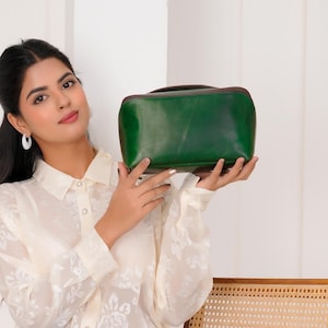 May include: A dark green leather handbag with a rectangular shape. The bag is held up by a person wearing a cream-colored floral blouse and blue jeans. The person is sitting on a wooden chair with a woven seat.