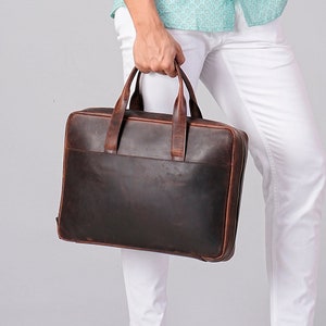 May include: A light green short-sleeved shirt with a subtle geometric pattern, paired with white trousers and trainers. A dark brown leather briefcase with top handles is held. Suitable for business or casual wear.