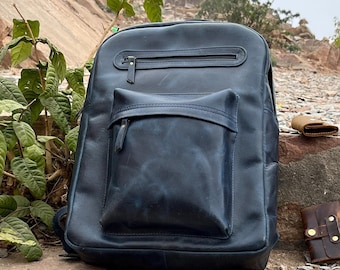 Könnte beinhalten: Ein marineblauer Lederrucksack mit einer Vordertasche und einem Reißverschluss oben. Der Rucksack steht auf einer Steinoberfläche mit einem kleinen braunen Portemonnaie, einem Leder-Notizbuch und einem Schlüsselbund.