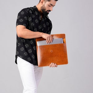 Könnte beinhalten: Eine braune Laptop-Hülle aus Leder mit schlichtem Design und einem einzelnen Knopfverschluss. Die Person trägt ein schwarzes Kurzarmhemd mit weißem Muster und eine weiße Hose. Der Hintergrund ist schlicht.