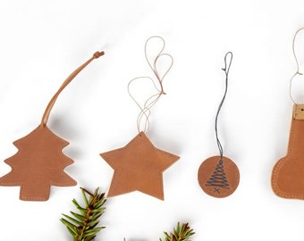May include: Four brown leather Christmas ornaments: a tree, a star, a circle with a tree design, and a stocking. The ornaments are hanging from strings on a white background.