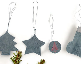 May include: Four gray leather Christmas ornaments with string hangers. The ornaments are a tree, a star, a ball with a red tree design, and a stocking.