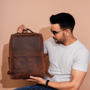 Puede incluir: Una mochila de cuero marrón con un acabado envejecido. La mochila tiene forma rectangular con un bolsillo frontal, bolsillos laterales y un asa superior. Cuenta con correas ajustables y cierre de cremallera. La mochila es sostenida por una persona.