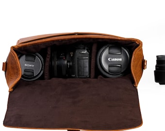 May include: A brown leather camera bag with a flap top and a buckle closure. The bag is open and has three compartments inside. The compartments contain a camera and two lenses. The lenses are labeled "Canon" and "Sony".