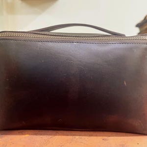 May include: A dark brown leather clutch bag with a top zip and a small handle. The rectangular bag has a smooth texture and a visible zip. The handle is attached to the top of the bag.