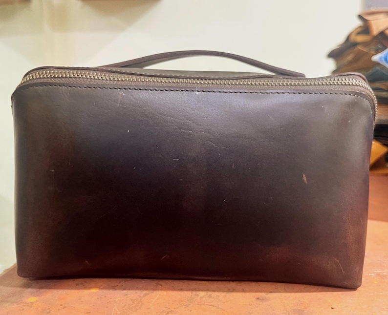 May include: A dark brown leather clutch bag with a top zip and a small handle. The rectangular bag has a smooth texture and a visible zip. The handle is attached to the top of the bag.