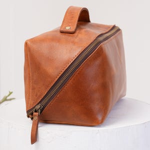 May include: A brown leather travel bag with a zipper closure and a top handle. The bag is made of a soft, supple leather and has a vintage look.