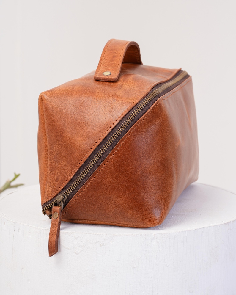 May include: A brown leather travel bag with a zipper closure and a top handle. The bag is made of a soft, supple leather and has a vintage look.