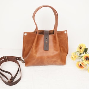 Könnte beinhalten: Eine braune Ledertasche mit einem braunen Lederriemen. Die Tasche hat einen oberen Griff und einen abnehmbaren Schulterriemen. Die Tasche befindet sich auf einer weißen Oberfläche.