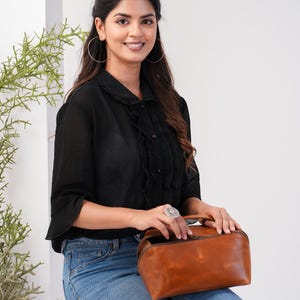 May include: A woman wearing a black blouse with a ruffled collar and blue jeans is holding a brown leather handbag with a zipper closure.
