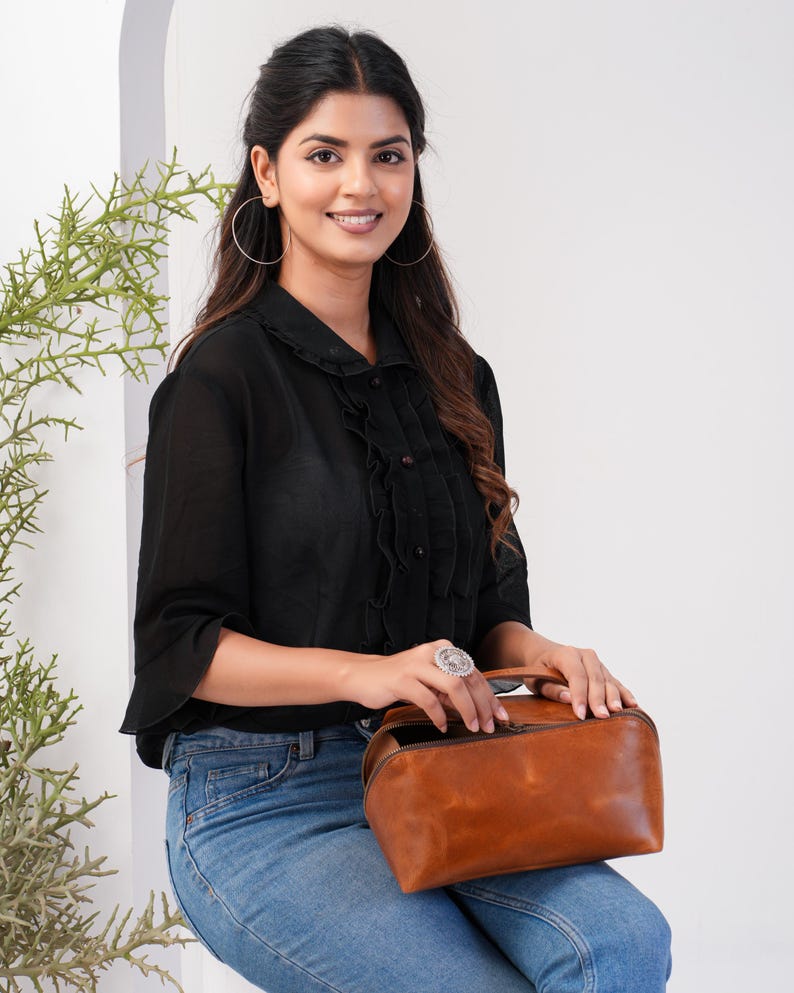 May include: A woman wearing a black blouse with a ruffled collar and blue jeans is holding a brown leather handbag with a zipper closure.