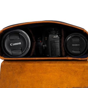 May include: Open brown leather camera bag with a Canon lens cap on the left and a Sony lens cap on the right. A black camera body is in the center. The bag has a blue interior.