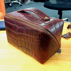 Puede incluir: Un neceser de viaje de cuero burdeos con un patrón de cocodrilo texturizado. El estuche rectangular tiene una cremallera envolvente, un asa superior y una cremallera dorada. Diseñado para viajes y almacenamiento.