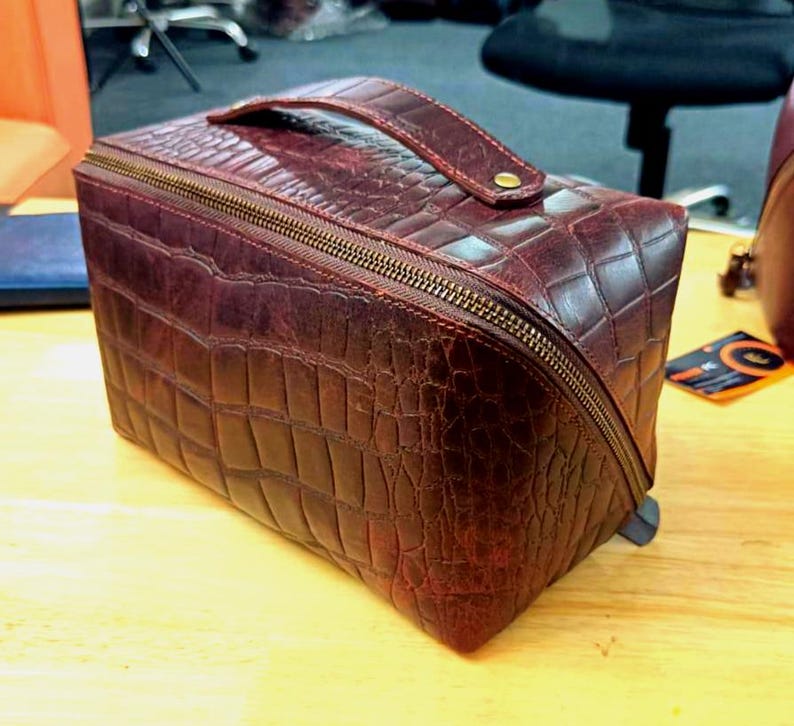 May include: A burgundy leather travel case with a crocodile skin effect. The rectangular case has a wraparound zip, a top handle, and a gold-toned zip. The case is designed for travel and storage.