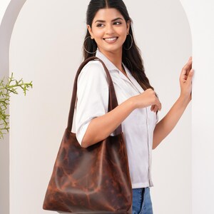 May include: A woman wearing a white shirt and blue jeans is holding a brown leather tote bag. The bag has a distressed look and is made of high-quality leather.