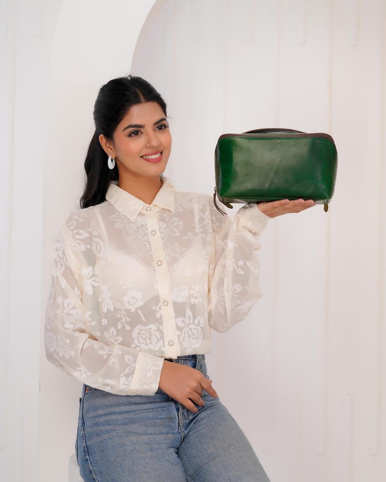 May include: A dark green leather rectangular bag with a brown trim and a zip closure. The bag is held in front of a person wearing a white floral blouse and blue jeans.