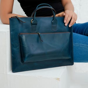 Op de afbeelding: Een teal leren tote bag met een handvat aan de bovenkant en een voorvak. De tas heeft een rechthoekige vorm met een ritssluiting en een bruine ritstrekker. De tas staat op een wit oppervlak.