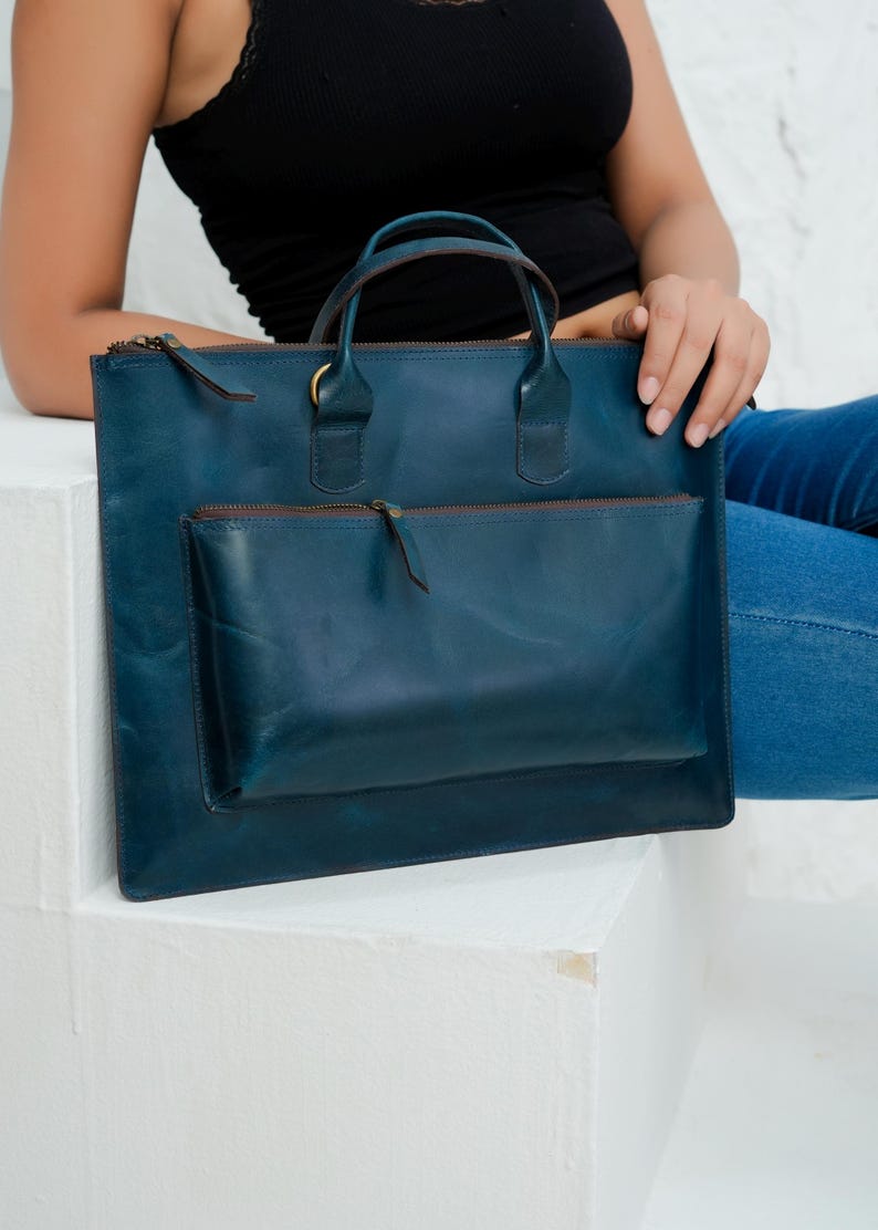 Op de afbeelding: Een teal leren tote bag met een handvat aan de bovenkant en een voorvak. De tas heeft een rechthoekige vorm met een ritssluiting en een bruine ritstrekker. De tas staat op een wit oppervlak.