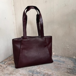 May include: A burgundy leather tote bag with two long handles. The bag has a rectangular shape with a structured design and a zipper closure. The bag is made of a smooth, solid-colored leather.