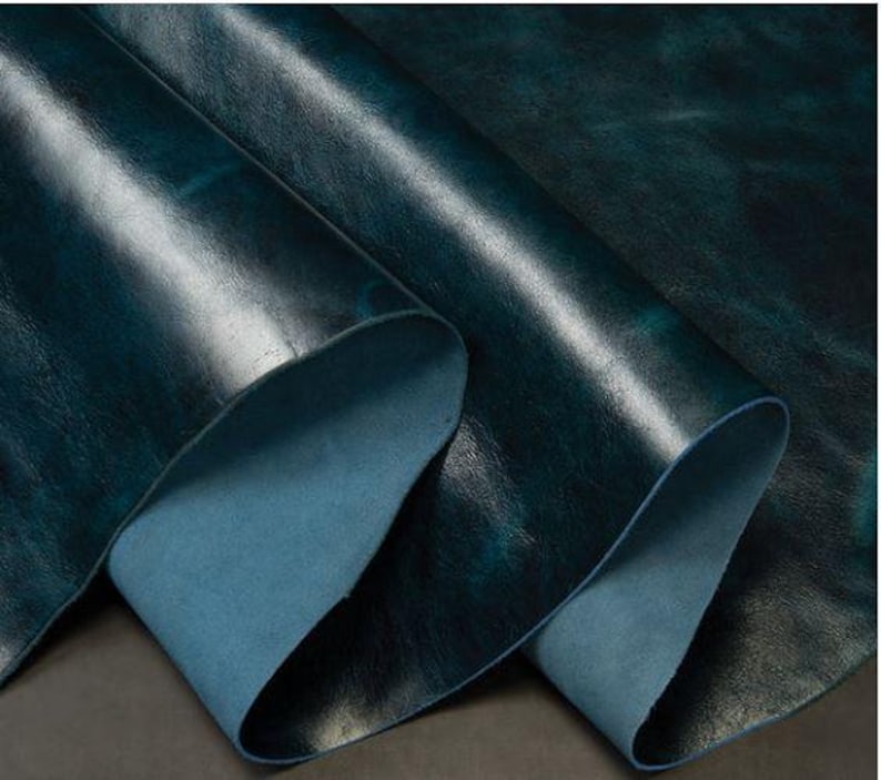 May include: Close-up of a deep teal leather with a textured, distressed finish. The leather is folded over, revealing a lighter blue underside.