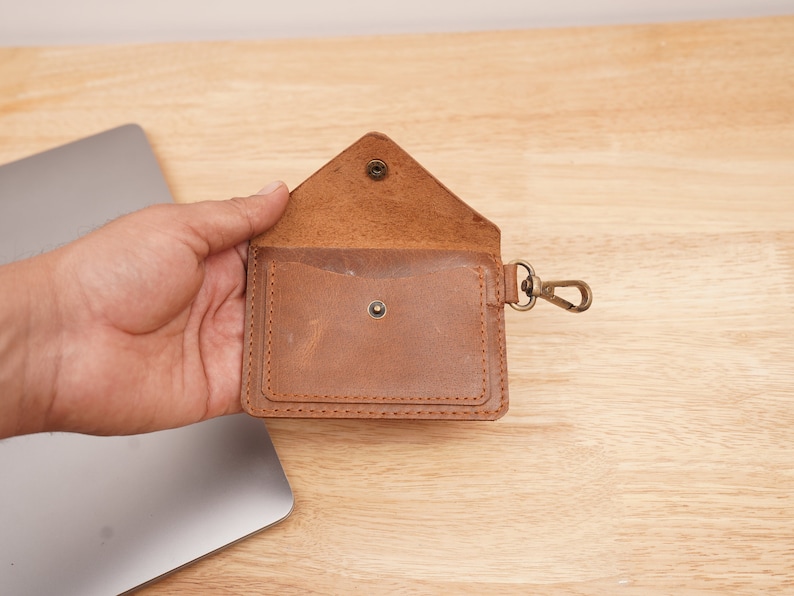 May include: A brown leather wallet with a flap closure and a metal clasp. The wallet has a small pocket on the front and a larger pocket on the inside. The wallet is attached to a metal key ring.