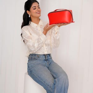 May include: A vibrant red rectangular handbag with a top handle and black trim. The bag is held up by a person wearing a cream floral blouse and blue jeans. The background is white.