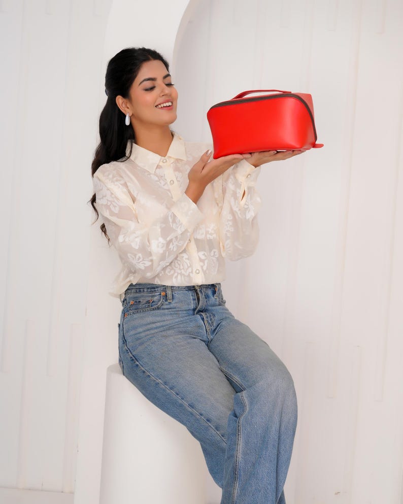 May include: A vibrant red rectangular handbag with a top handle and black trim. The bag is held up by a person wearing a cream floral blouse and blue jeans. The background is white.
