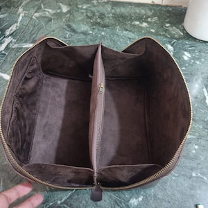 May include: Open brown leather travel bag with a zippered closure. The interior is divided into two compartments with a central zippered pocket. The bag has a soft, suede-like lining and a gold-coloured zipper.