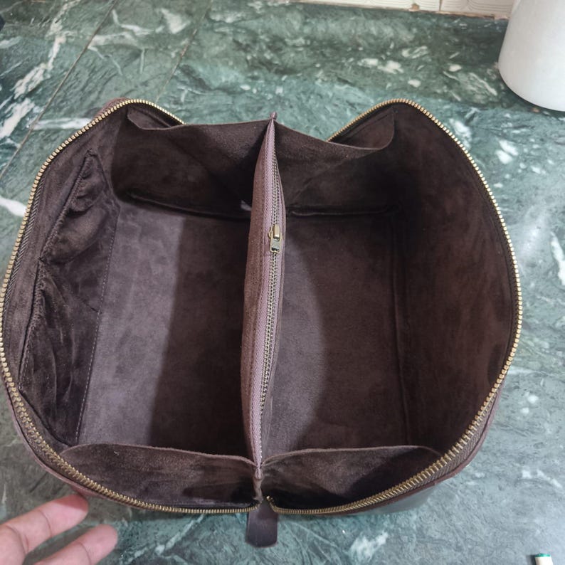 May include: Open brown leather travel bag with a zippered closure. The interior is divided into two compartments with a central zippered pocket. The bag has a soft, suede-like lining and a gold-coloured zipper.