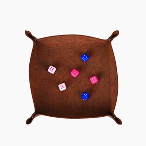 May include: A brown leather dice tray with rounded corners and four loops. Six multi-colored dice are scattered inside the tray. The dice are white, pink, and blue with white pips.