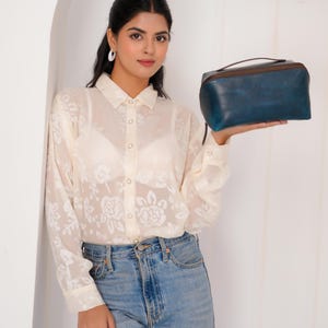 May include: A blue leather travel bag with a brown zip and handle. The bag is rectangular with rounded edges. The bag is held by a person wearing a white floral blouse and blue jeans.