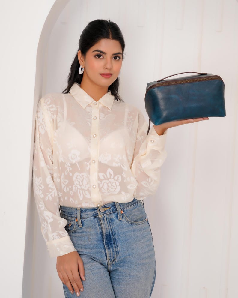 May include: A blue leather travel bag with a brown zip and handle. The bag is rectangular with rounded edges. The bag is held by a person wearing a white floral blouse and blue jeans.