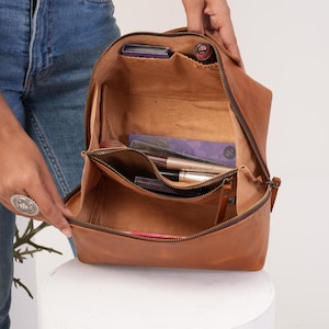 May include: Open tan leather cosmetic bag with a zip. The bag is filled with makeup items, including lipsticks, eyeshadow palettes, and makeup brushes. The bag is held open, revealing the interior compartments and contents.