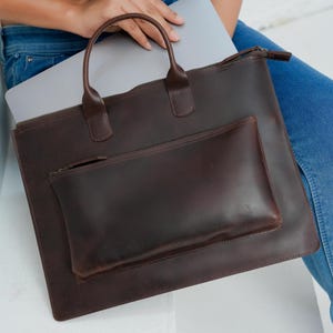 Op de afbeelding: Een donkerbruine leren laptoptas met een handvat en een kleinere zak aan de voorkant. De tas heeft een ritssluiting en is ontworpen om een laptop te dragen. De tas heeft een rijke, effen kleur.
