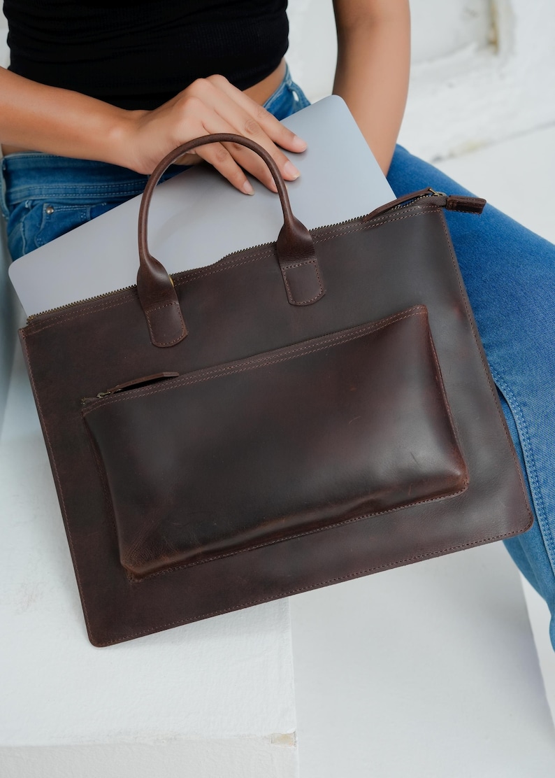 Op de afbeelding: Een donkerbruine leren laptoptas met een handvat en een kleinere zak aan de voorkant. De tas heeft een ritssluiting en is ontworpen om een laptop te dragen. De tas heeft een rijke, effen kleur.