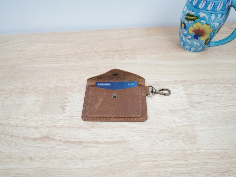 May include: A brown leather wallet with a card slot and a metal key ring. The wallet is open and a blue card is visible inside.